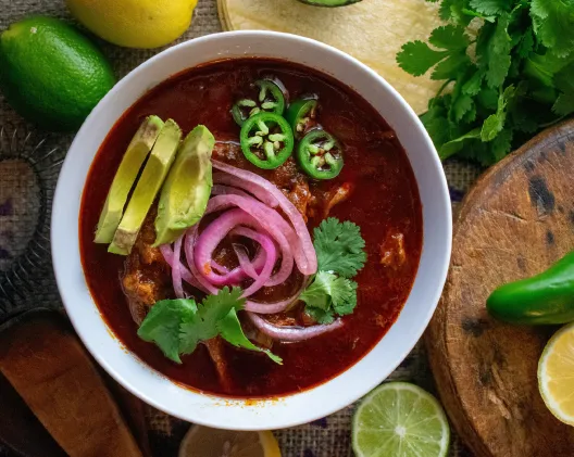 Bowl of birria in rich red broth garnished with avocado, red onion, cilantro, and jalapeno slices