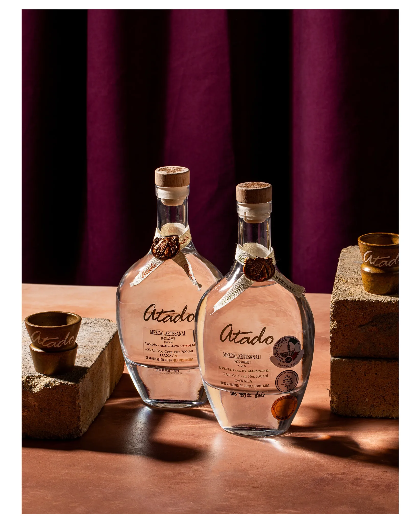 Espadín and Tepeztate bottles with branded copitas on adobe bricks, velvet curtain backdrop