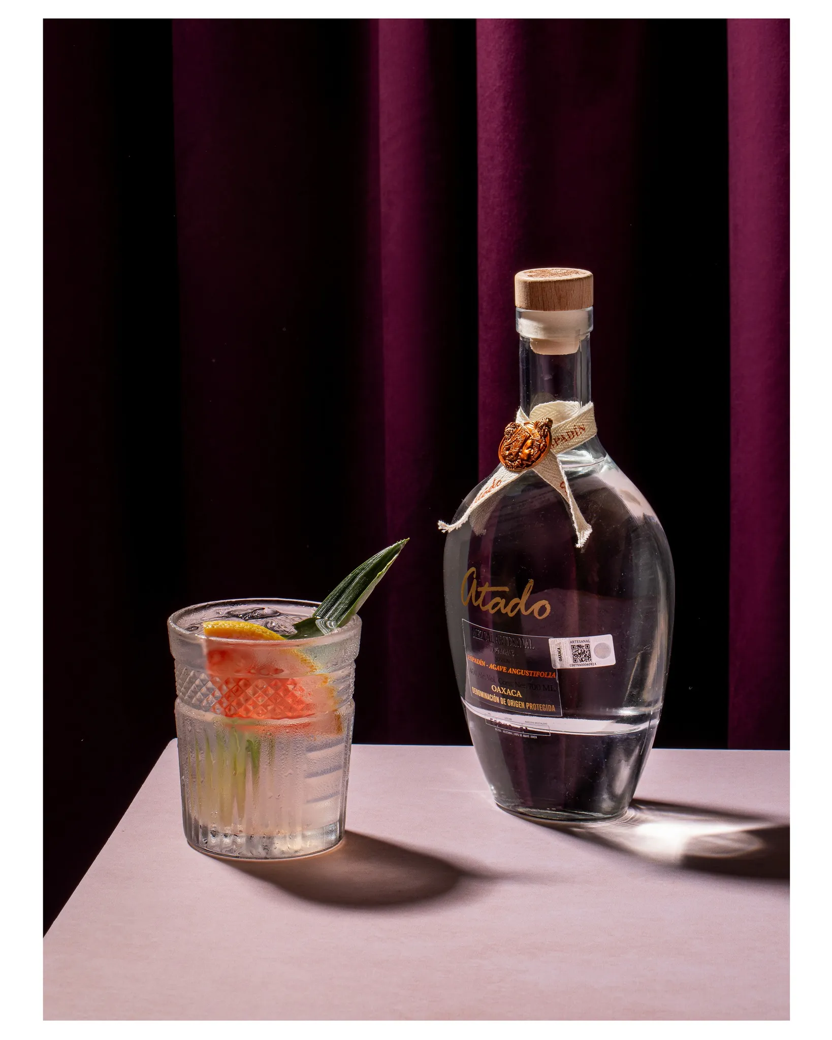 Espadín bottle with citrus mezcal cocktail against dark velvet curtain