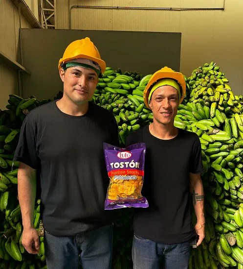 Two factory workers in hard hats holding LuLu Toston bag in front of plantain bunches