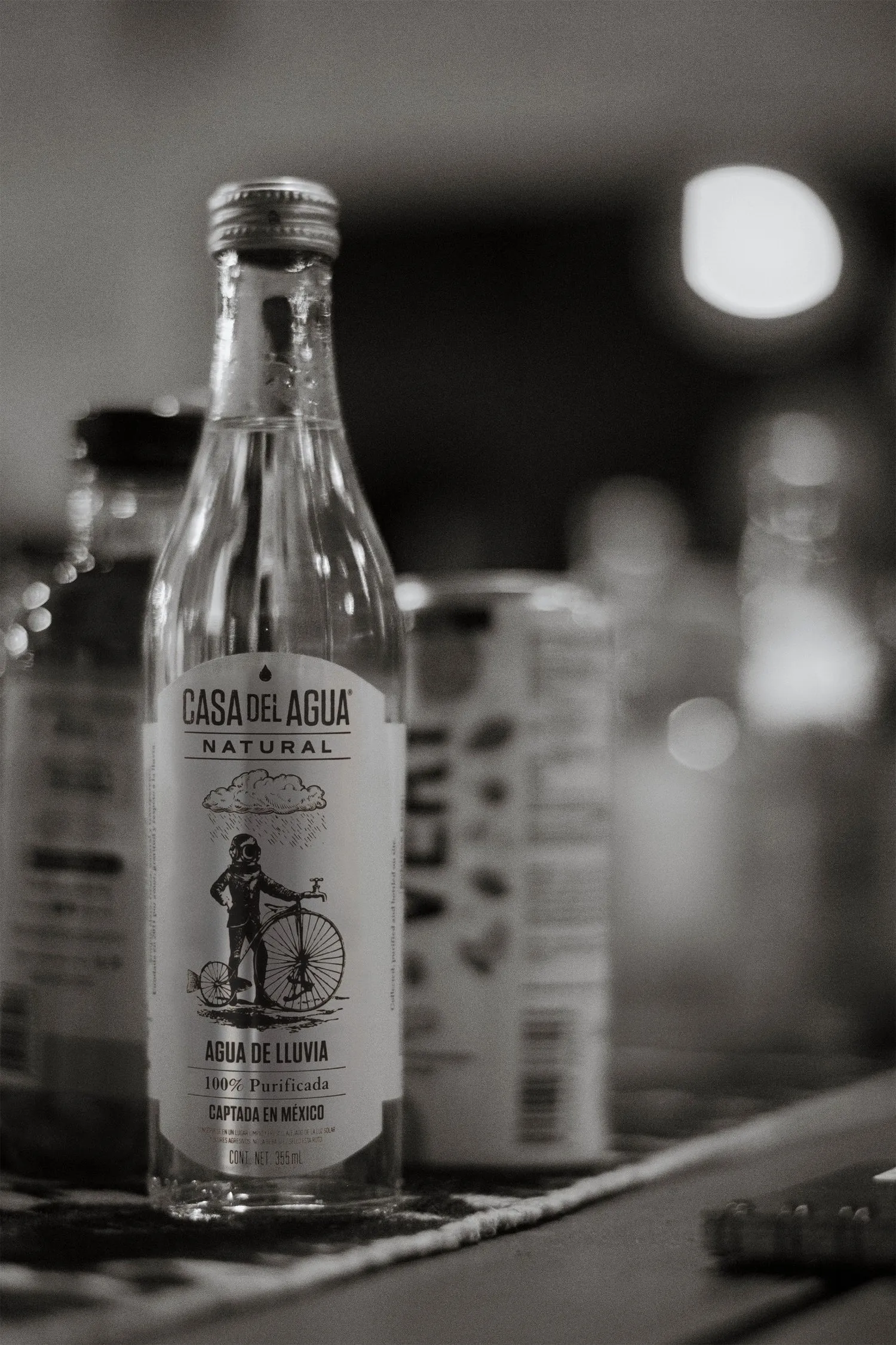 Black-and-white photo of Casa del Agua Natural bottle on a bar counter with bokeh lights