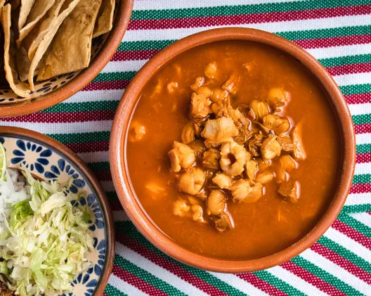 Bowl of pozole rojo with hominy and pork on a colorful Mexican serape with shredded cabbage and tostadas