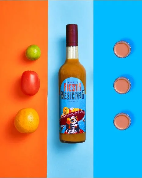 Sangrita bottle on orange and blue backdrop with citrus and shot glasses