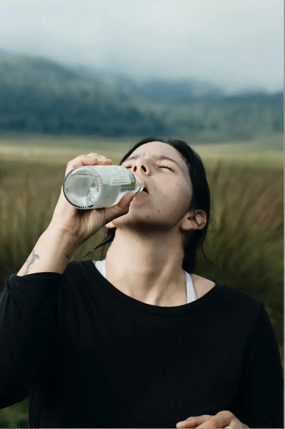 Woman drinking from a Casa del Agua bottle in a grassy field with mountains in the background