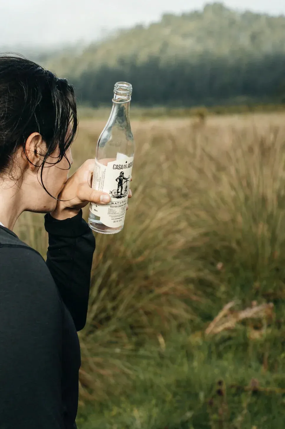 Woman seen from behind holding a Casa del Agua bottle while standing in a grassy field