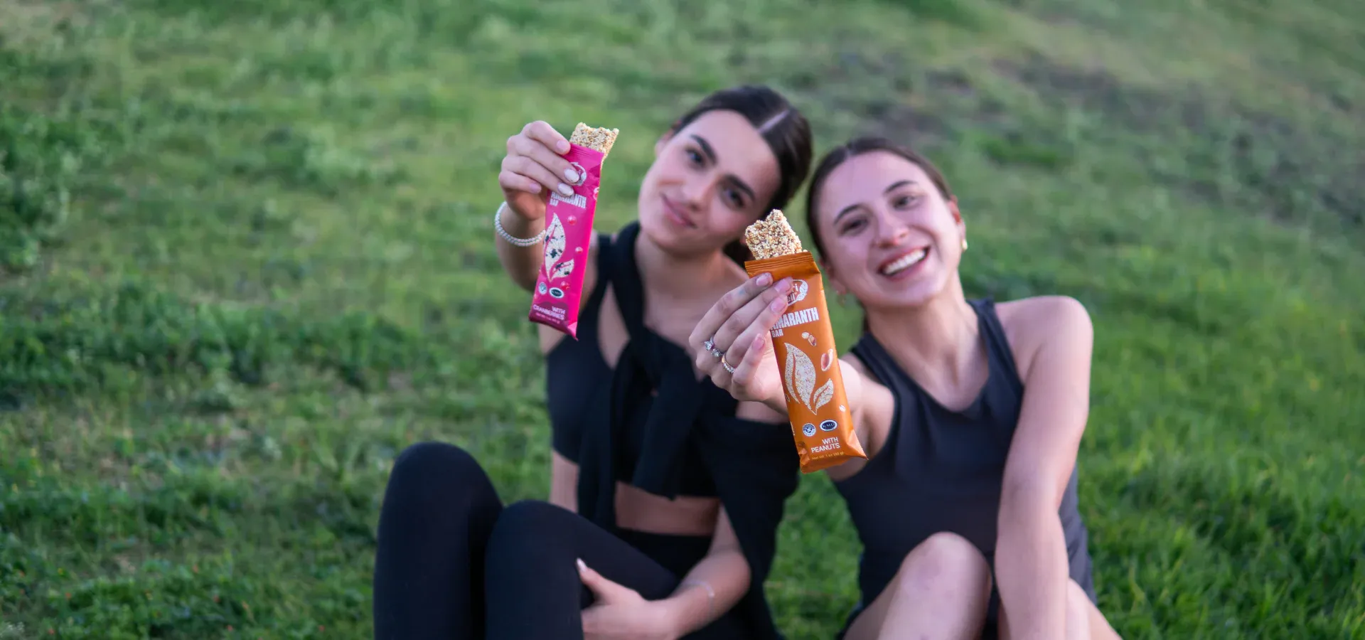 Two women on grass holding Nutri Well amaranth bars in landscape format